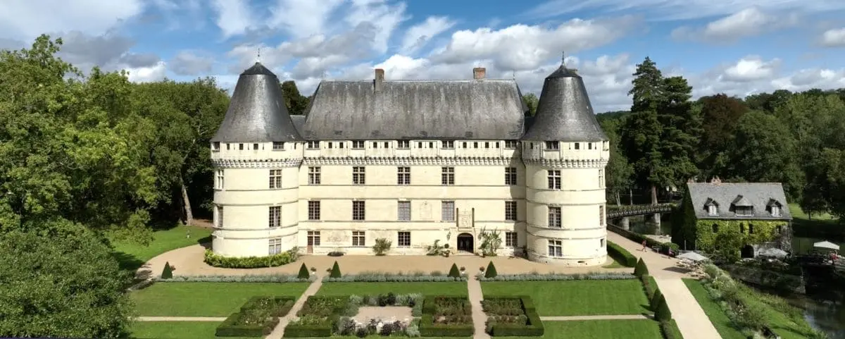A picturesque French castle with two large, round towers and a central building, nestled in a green landscape with a walking path leading to the entrance.