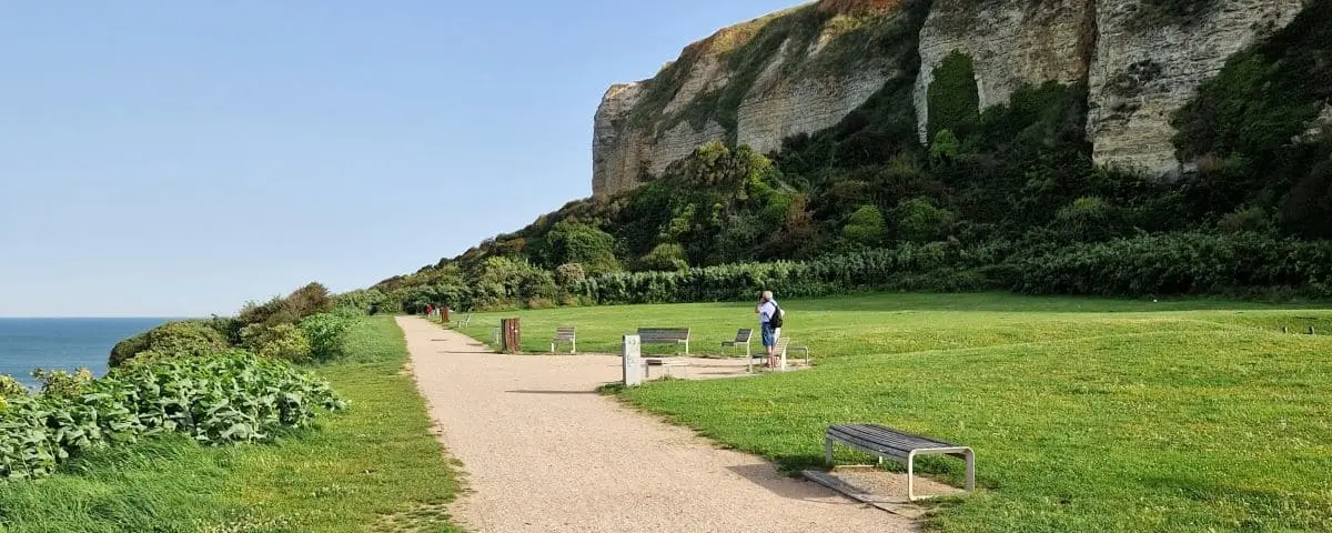 A scenic view of Jardin des Hauteurs des Falaises in Le Havre, featuring a gravel path, green lawns, and a backdrop of tall, rocky cliffs near the sea.