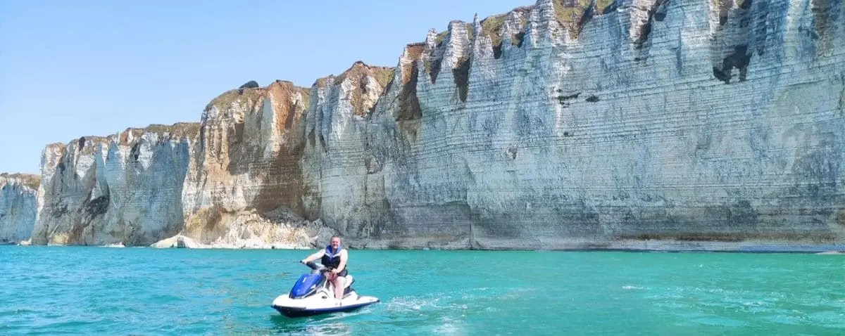 A person riding a jet ski near the towering white cliffs of Fécamp, France, with clear blue water and the dramatic cliffs in the background.