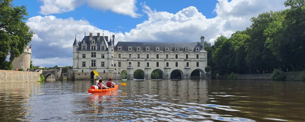 A group of people kayaking on the Cher River with the Château de Chenonceau in the background. The castle's arches span the river, and the scene is set under a partly cloudy sky.
