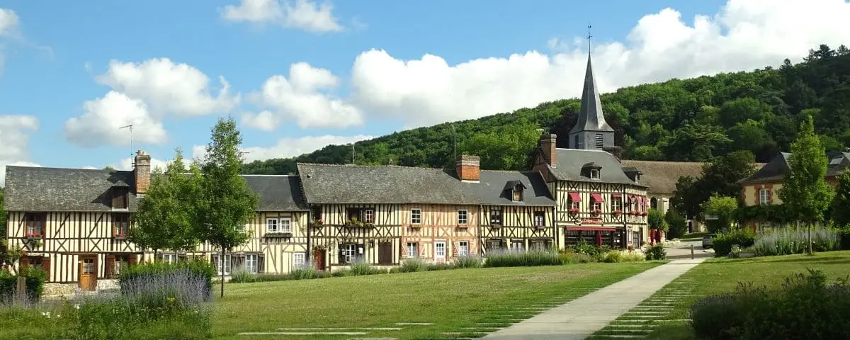 Scenic view of Le Bec-Hellouin village with charming half-timbered houses, a church with a spire, and a well-manicured lawn under a partly cloudy sky.