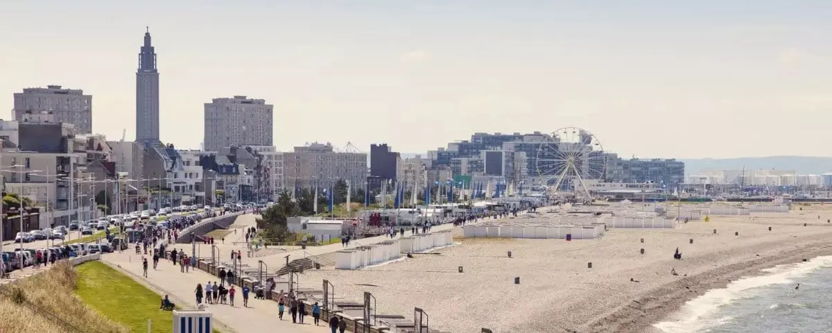 Coastal view of Le Havre city, highlighting a sandy beach, a promenade filled with people, and a Ferris wheel amidst modern buildings and skyscrapers.