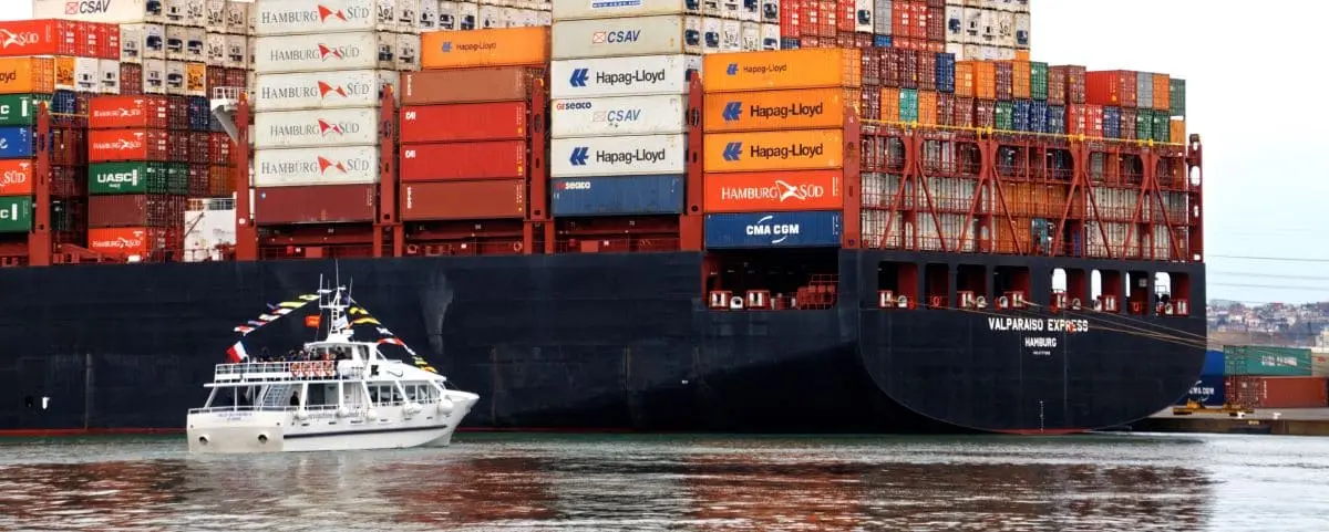 A small tour boat adorned with flags sails past a massive container in Le Havre Port.