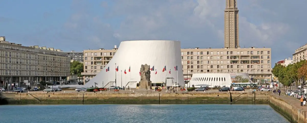 A view of Le Volcan, a modern white building with a curved design, located in Le Havre, surrounded by water and flags.