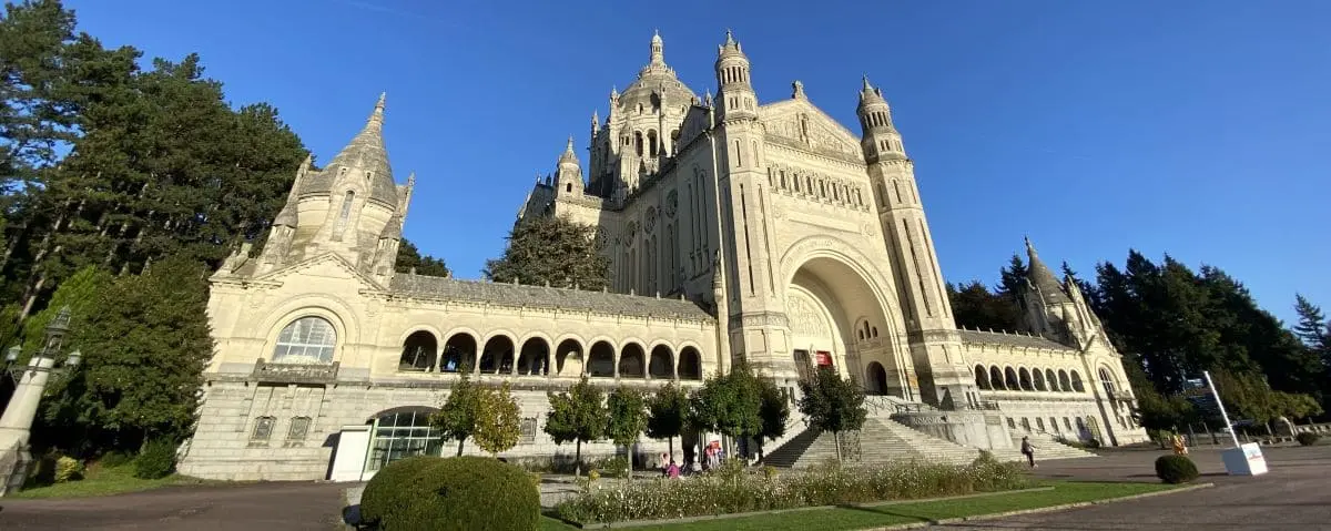 The grand Lisieux Basilica with its ornate façade, arched entrances, and spires, set against a clear blue sky and framed by trees.