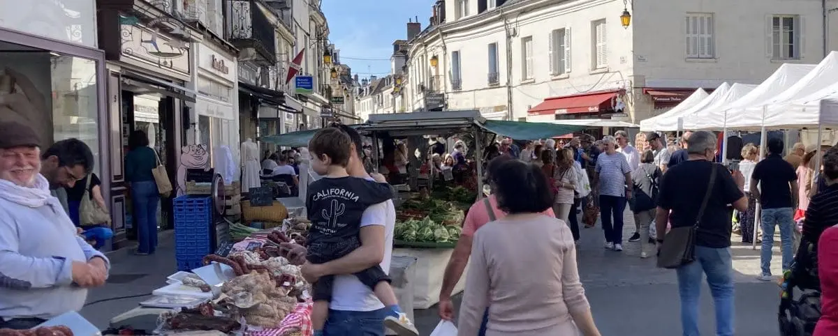 A bustling outdoor food market in Loches, filled with stalls offering various goods. Shoppers walk through the market, engaging with vendors and examining fresh produce.
