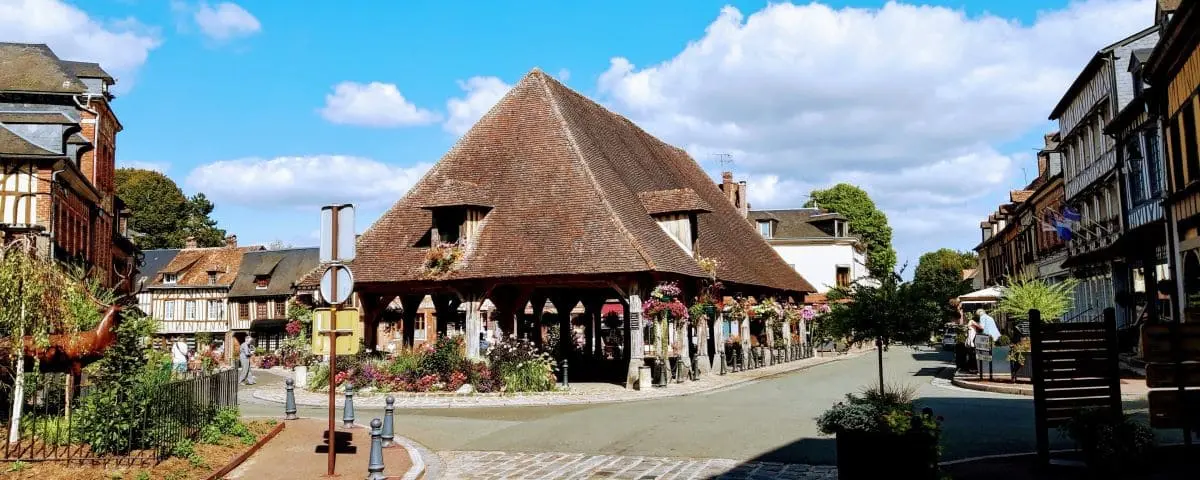 Charming village scene of Lyon-la-Forêt with half-timbered houses and a large, thatched market hall in the center, surrounded by a cobblestone street and flowers.