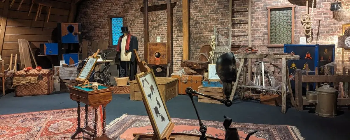 An exhibit filled with magical artifacts, including a guillotine, costumes, and various props used in magic shows, set in a rustic room with brick walls.