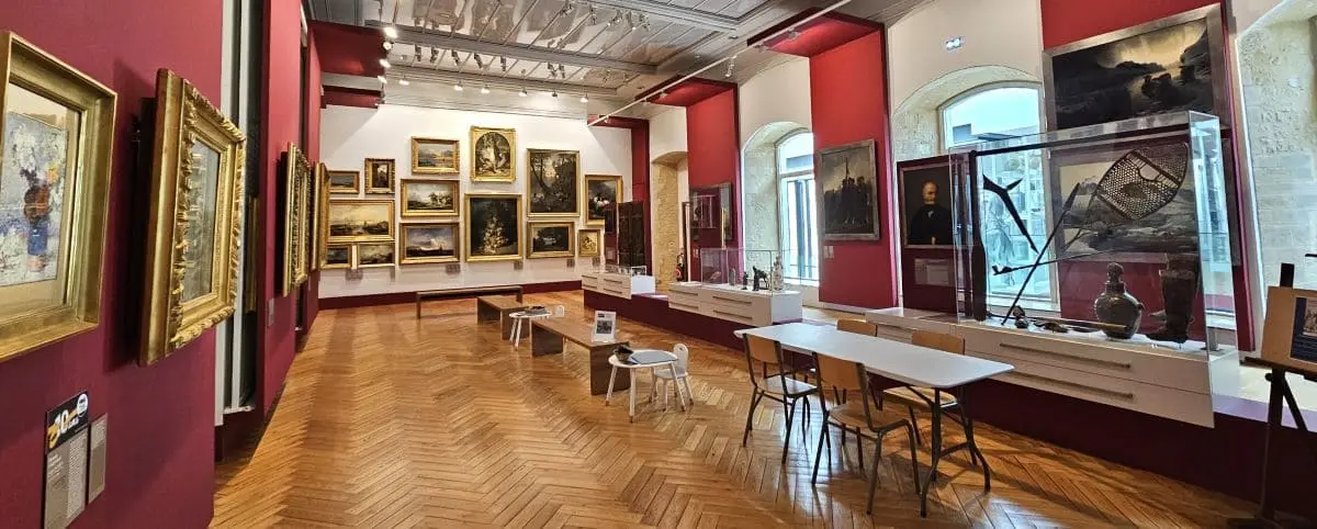 An art gallery room in the MAHB Museum featuring a variety of framed paintings on the walls, display cases with artifacts, and wooden parquet flooring.