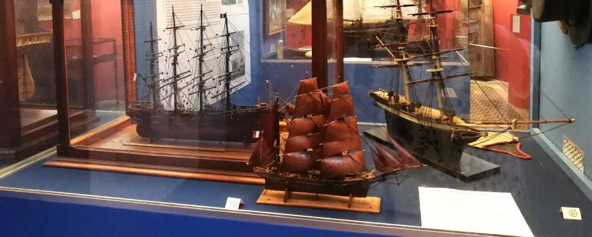 A display case with detailed ship models, including a tall ship with red sails and another with black sails. The museum showcases the rich maritime history of Honfleur.