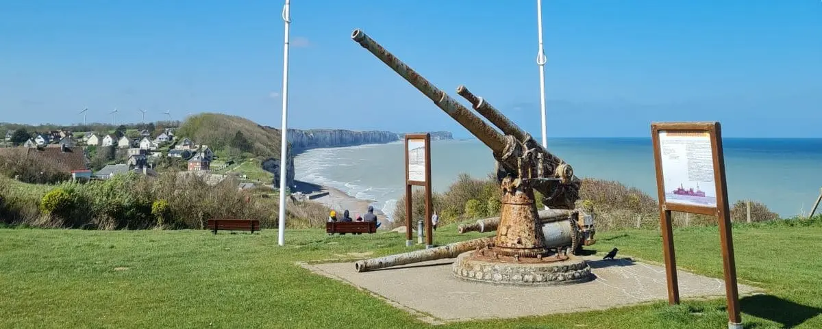 A war memorial featuring a large, rusted cannon with informational plaques on either side. The backdrop includes green lawns, a coastline, and distant cliffs.