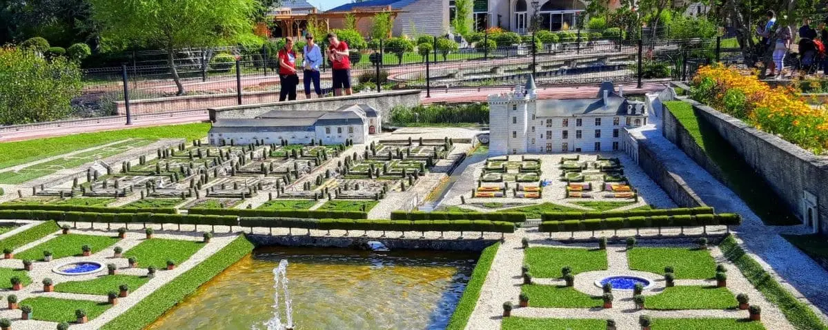 A detailed miniature replica of a castle and its surrounding gardens, displayed in an outdoor park. The scene includes a small pond with a fountain, well-trimmed hedges, and visitors observing the intricate model.