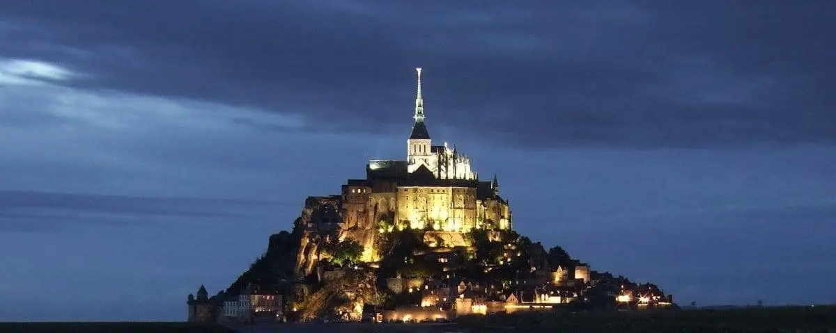 Mont Saint-Michel illuminated at night, with the abbey and surrounding buildings glowing against the dark sky.