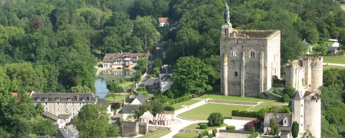 A medieval fortress with a tall, square keep and surrounding walls, situated on a hill with trees and a small village visible in the background.