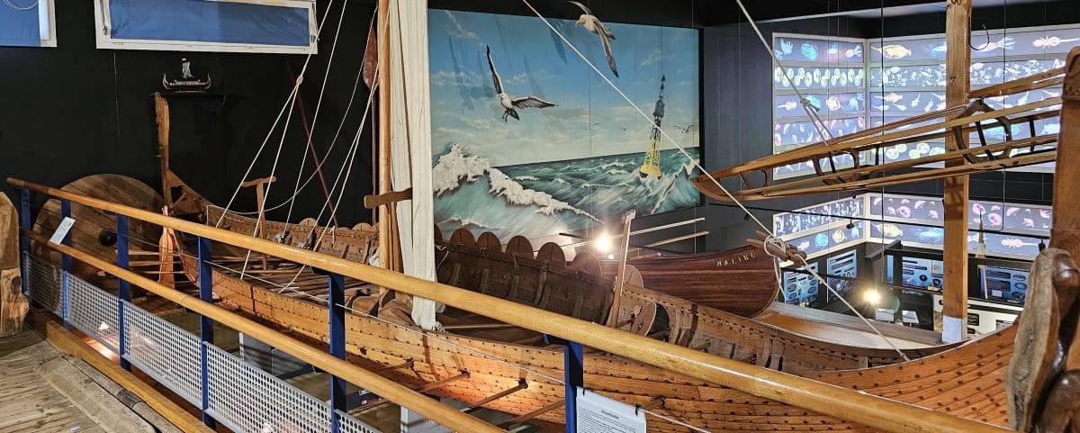 A detailed model of a wooden ship displayed inside the Estran Museum in Dieppe, with a backdrop of a painted ocean scene and various maritime exhibits.