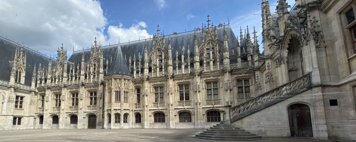 The ornate facade of the Normandy Parliament building in Rouen, featuring intricate Gothic architecture, spires, and detailed stone carvings under a partly cloudy sky.