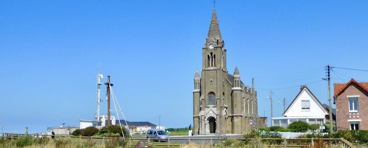 The Notre-Dame de Bonsecours Chapel in Dieppe, featuring gothic architecture with a tall spire, situated near residential houses and a clear blue sky.