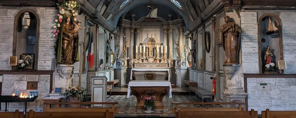 The interior of Notre Dame de Grace Chapel in Honfleur, featuring an ornate altar with multiple candles and religious statues. The chapel's walls are adorned with plaques and floral arrangements.