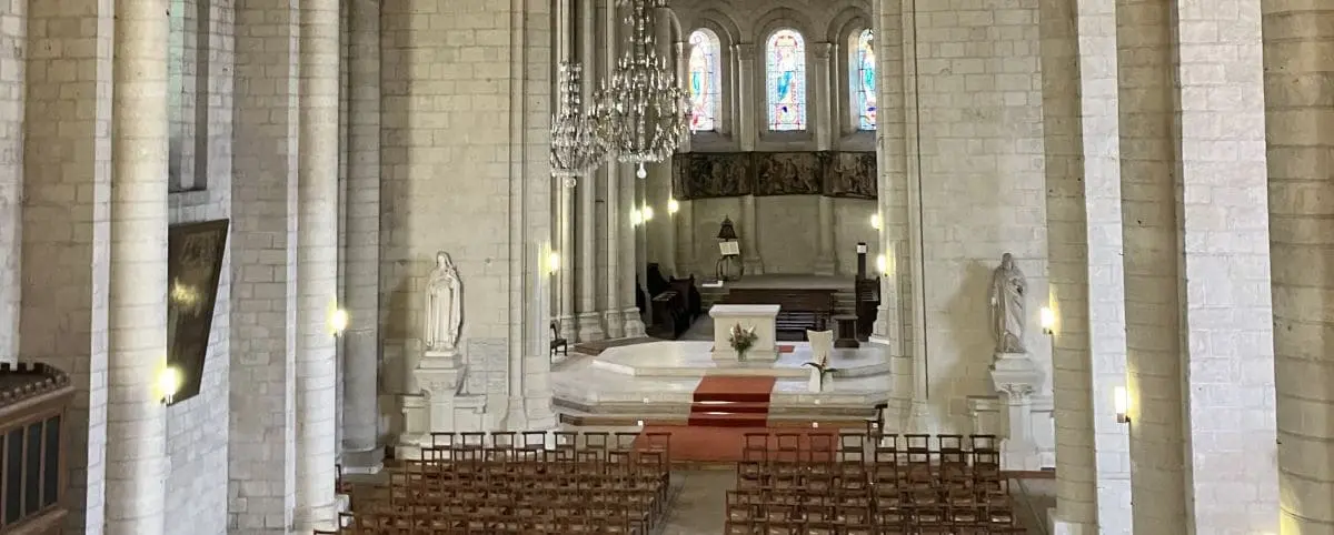 Inside Notre-Dame de Nantilly Church in Saumur, featuring a spacious nave with rows of wooden chairs, ornate chandeliers, and stained glass windows.