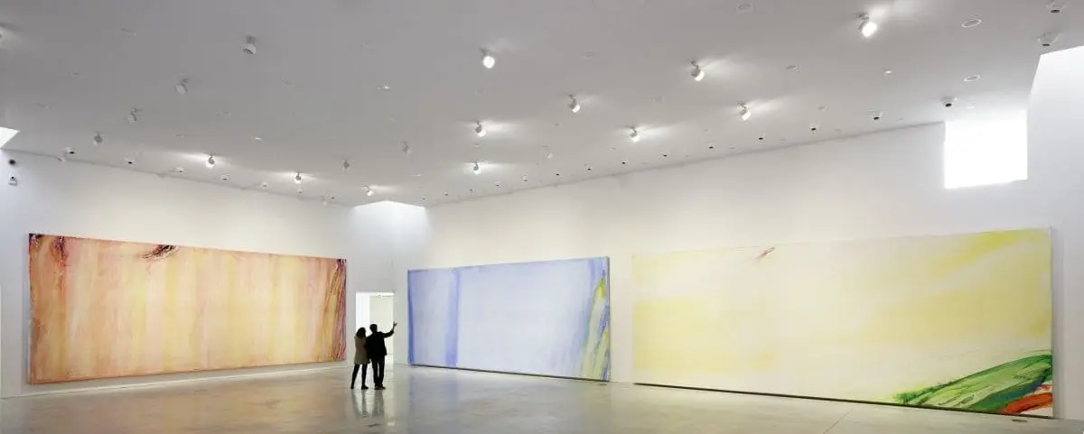 A spacious art gallery in the Olivier Debré Center in Tours, France, with high ceilings, displaying large abstract paintings on white walls, with two people observing the artwork.