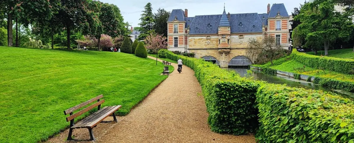 A peaceful park with a gravel path leading to a historic building with a moat. The path is lined with green hedges, benches, and lush grass, creating a serene atmosphere.
