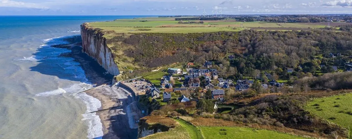 An aerial view of Petites Dalles, featuring dramatic cliffs beside a coastal village, with the sea waves gently hitting the shore and wind turbines visible in the distance.