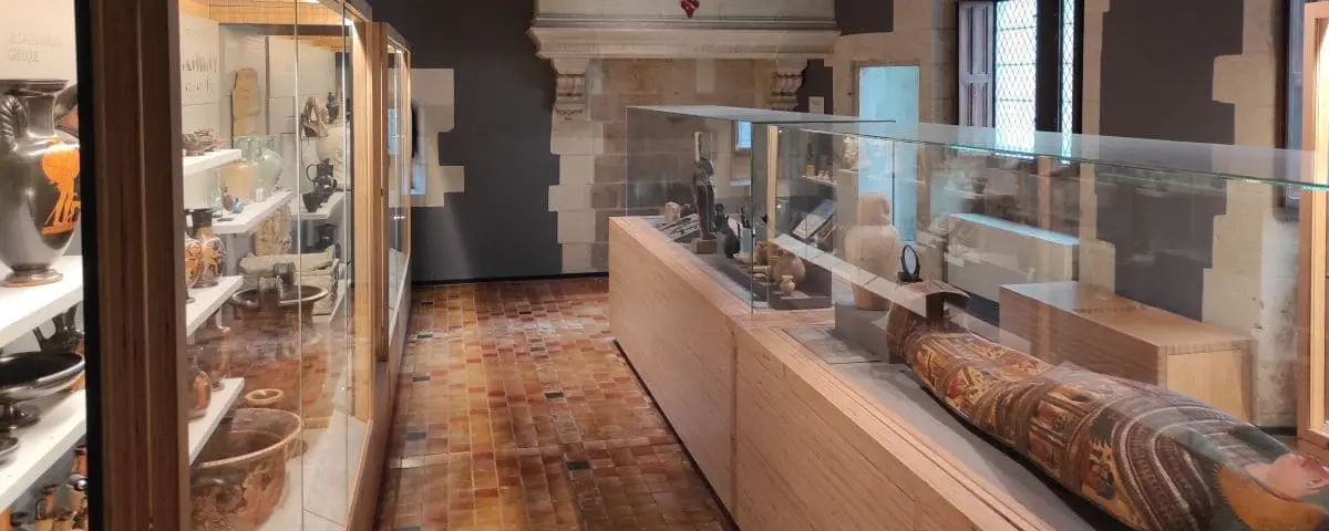 Exhibit room at the Pincé Museum displaying ancient artifacts, pottery, and an Egyptian mummy in glass cases, with tiled flooring and stone walls.