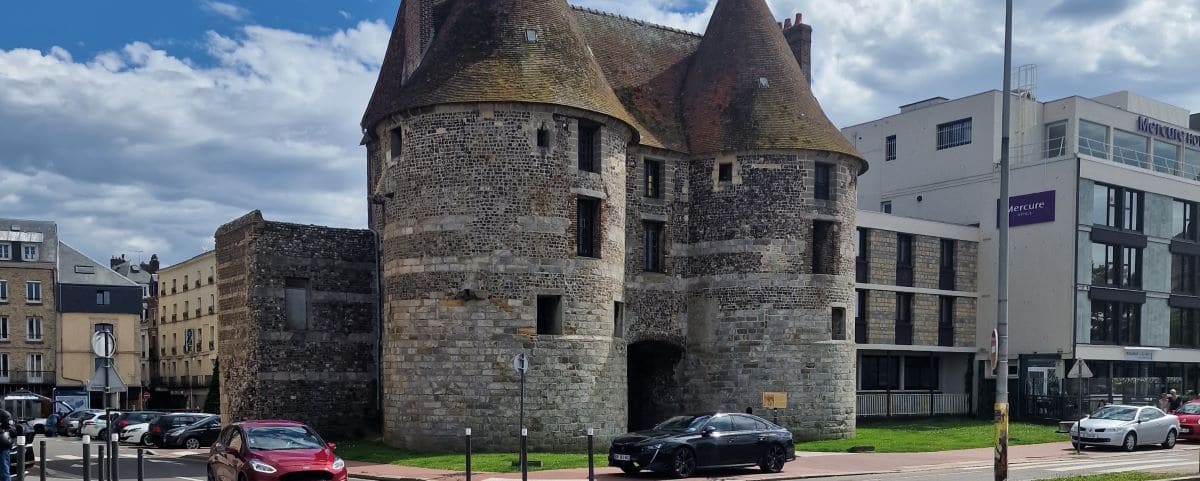 The Porte des Tourelles, an ancient stone gateway with round towers, located in Dieppe, surrounded by modern buildings and parked cars.