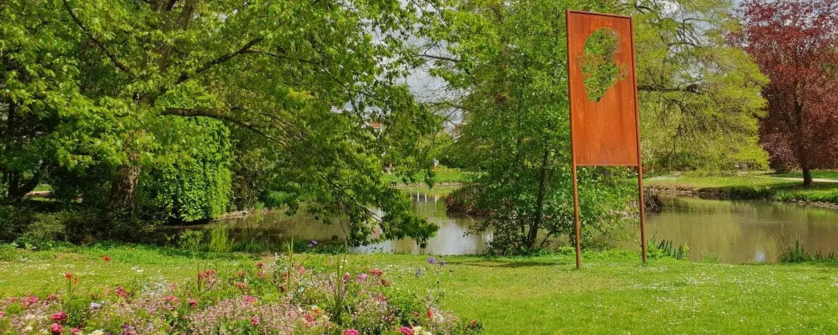 A tranquil garden in Tours, France, with vibrant flowers, lush trees, and a small pond, featuring a unique metal sculpture with a heart-shaped cutout.