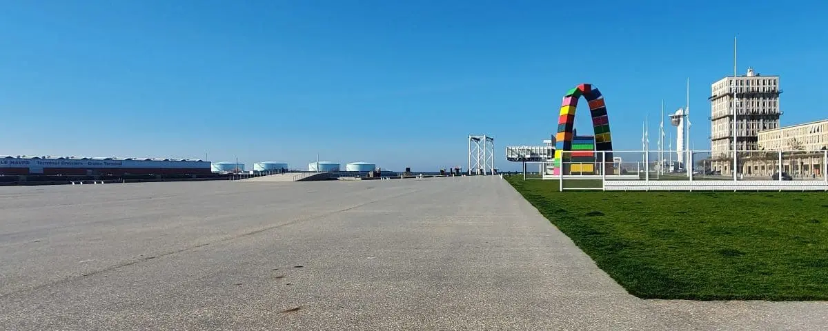 The wide expanse of the quay in Le Havre, featuring a large colorful arch sculpture and industrial structures in the background.