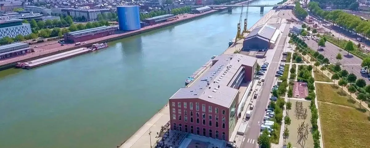 An aerial view of the quays along the Seine River in Rouen, with modern buildings, green spaces, and industrial areas lining the waterfront, and boats docked along the river.