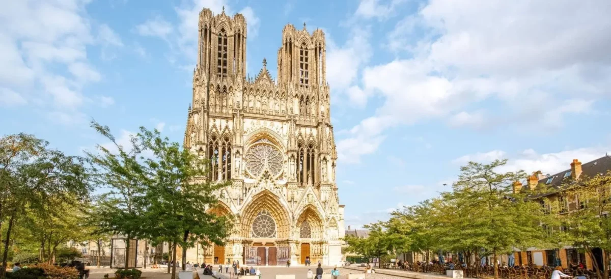 Reims Cathedral, a masterpiece of Gothic architecture, stands under a clear blue sky, flanked by green trees with people milling about the square.