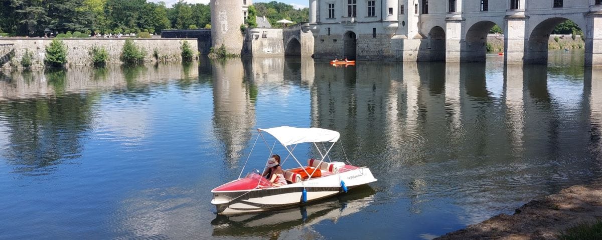 A small, white rental boat with a canopy cruising on the calm waters of the Cher River near the Château de Chenonceau. The scene is peaceful with clear reflections on the water.
