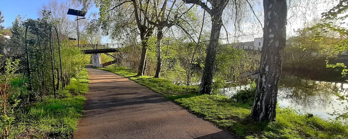 A peaceful path along the riverbanks in Périgueux, bordered by trees and greenery. The scene captures a quiet, natural setting perfect for walking or cycling, with a small bridge visible in the background.