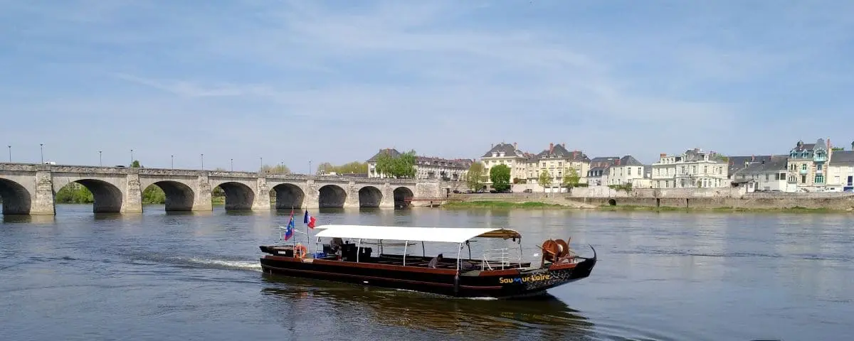 A traditional boat cruising along the river in Saumur, with a historic stone bridge and charming buildings in the background.