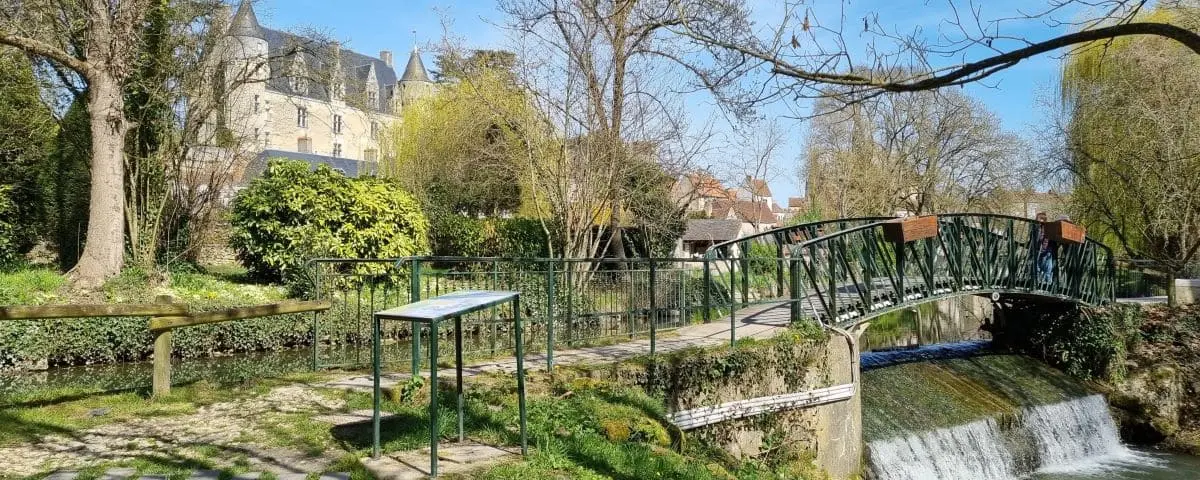 A serene riverfront view in Montrésor with a small bridge, lush vegetation, and the castle in the background, bathed in sunlight.