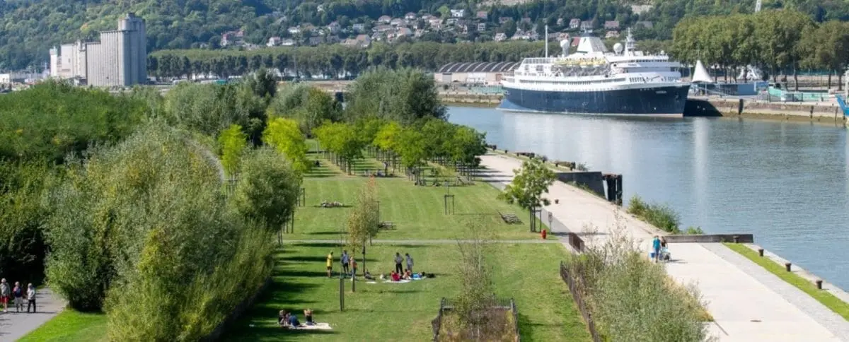 A lush, green park area in Rouen, known as Rollet Park, with people enjoying the open space, trees lining the pathways, and a large ship docked along the Seine River.
