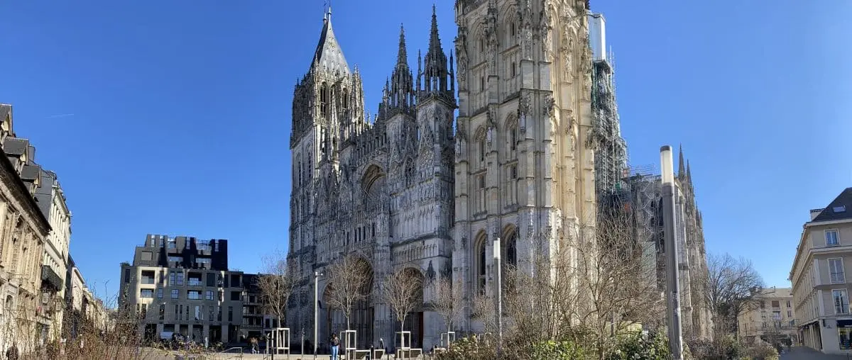 The impressive Rouen Cathedral, showcasing its towering Gothic spires and elaborate facade, set against a clear blue sky with surrounding historic buildings.