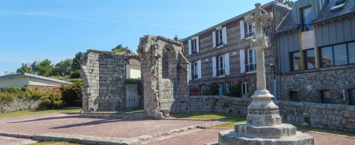 Ruins of the Saint Nicolas Church with partially standing stone walls and a cross in the courtyard. Modern buildings are visible in the background.