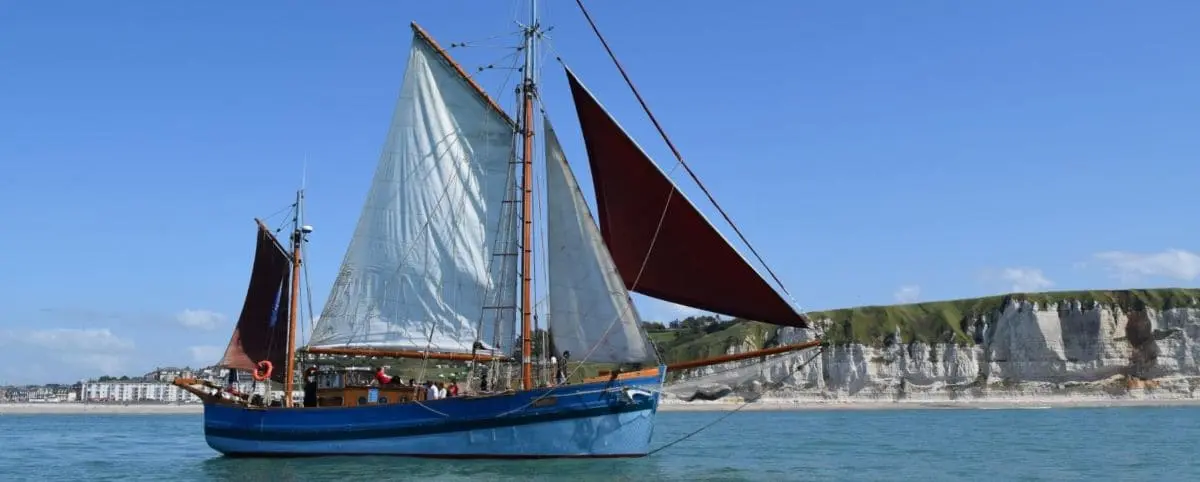 A traditional sailing boat with white and red sails on the waters near Fécamp, with the town and cliffs visible in the background.