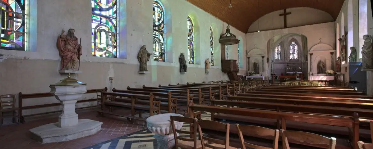 The interior of Saint-André Church, featuring wooden pews, statues, and stained glass windows, with light streaming in and illuminating the serene space.