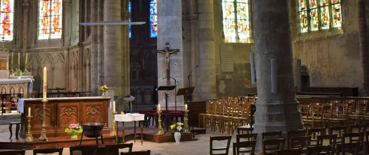 The interior of Saint Brice Church featuring a detailed wooden altar with candles and floral arrangements. The space is illuminated by colorful stained glass windows, and rows of wooden chairs are arranged for the congregation.