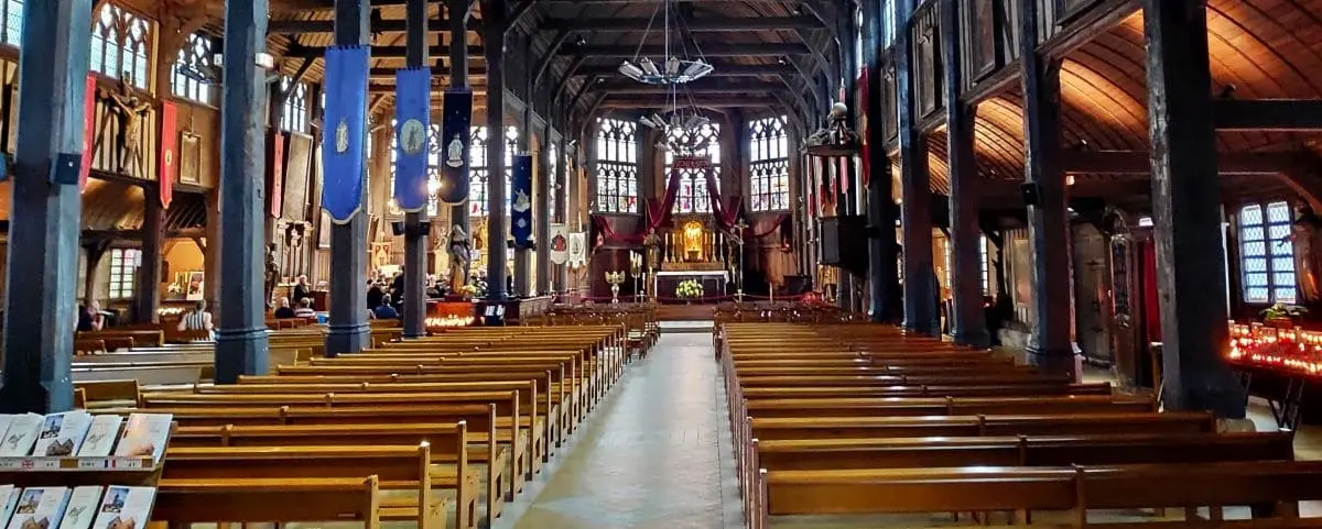 A spacious interior of Saint Catherine Church, showcasing wooden beams, stained glass windows, and banners hanging from the ceiling. Rows of wooden pews lead up to the altar.