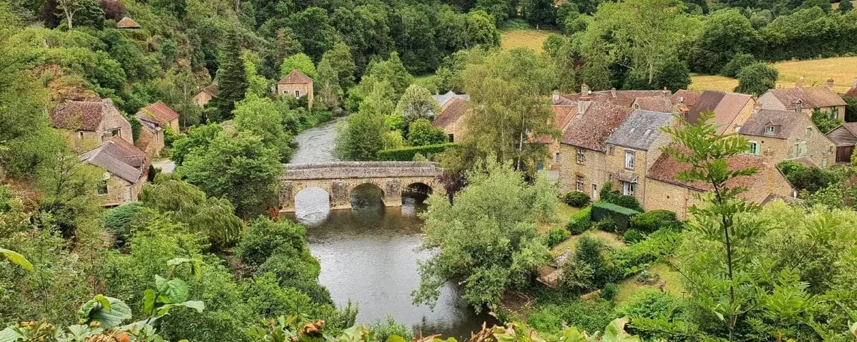 Tranquil scene of Saint-Ceneri-le-Gerei, featuring a stone bridge over a river, surrounded by quaint houses and lush greenery.