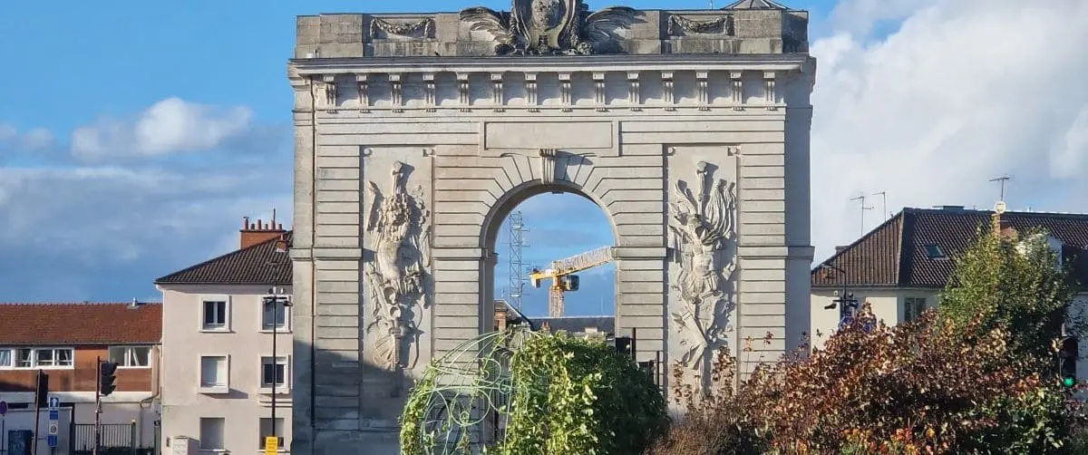 The Saint Croix Gate, an ornate stone arch with sculpted reliefs, stands prominently against a blue sky. Surrounding the gate are residential buildings and greenery.