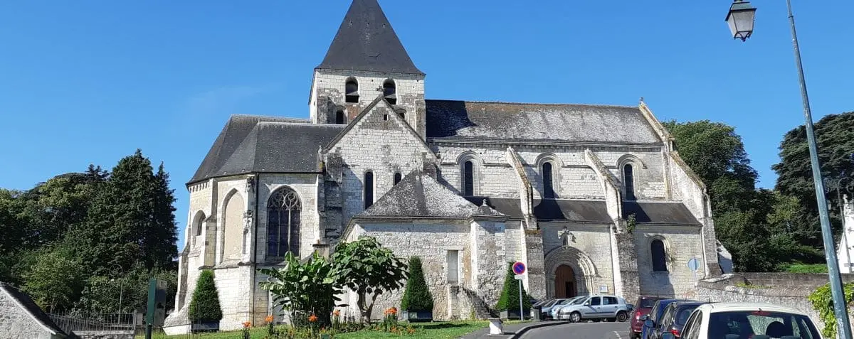 A beautiful stone church with a tall, pointed steeple set against a clear blue sky. The church is surrounded by lush greenery and parked cars, highlighting its historic and serene setting.