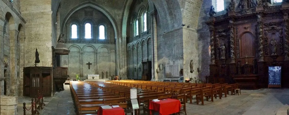 The interior of Saint-Etienne Church in Périgueux, showcasing its stone arches, wooden pews, and a simple altar with a cross. The high ceilings and stained glass windows add to the serene and historic ambiance.