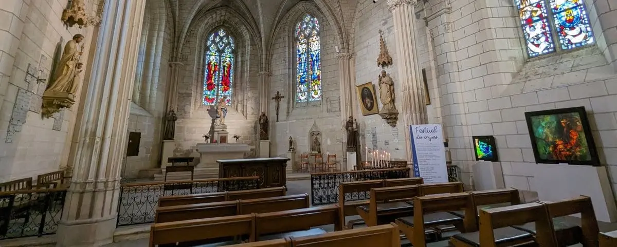 The interior of Saint Florentin Church, showcasing its Gothic architecture with high vaulted ceilings, stained glass windows, and wooden pews. The altar area is adorned with religious statues and vibrant, colorful windows.