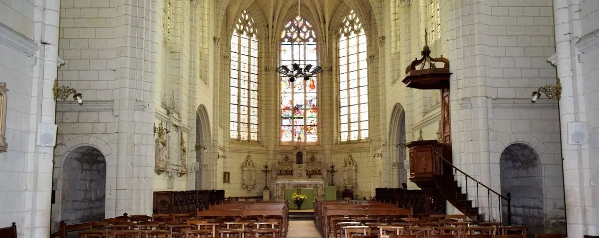 The interior of Saint-Jean-Baptiste Church in Montrésor, featuring a grand altar, high vaulted ceilings, and rows of wooden pews, with a large stained glass window at the back.