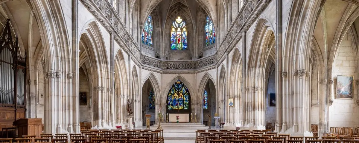 The interior of Saint Jean Church in Caen, showcasing its gothic architecture with tall arches and intricate stained glass windows. Rows of wooden chairs are arranged facing the altar.
